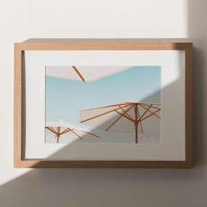 May include: A framed photograph of three white beach umbrellas against a blue sky. The umbrellas are made of fabric and have wooden frames. The photograph is in a light wood frame.