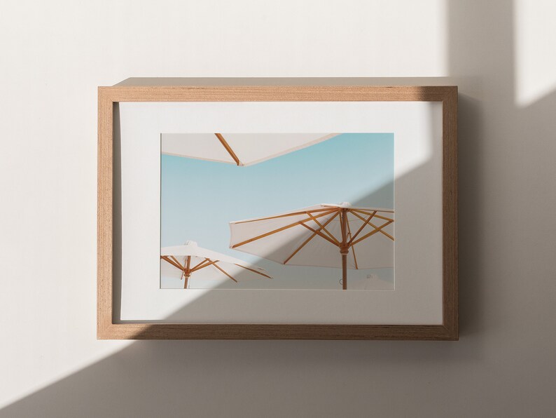 May include: A framed photograph of three white beach umbrellas against a blue sky. The umbrellas are made of fabric and have wooden frames. The photograph is in a light wood frame.