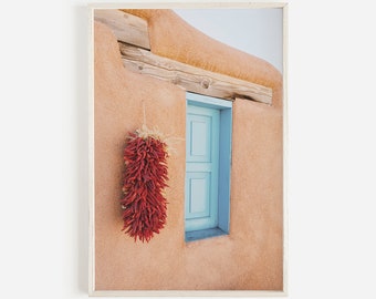 Puede incluir: Una ristra de chiles rojos cuelga fuera de una ventana de un edificio con paredes de adobe. La ventana está pintada de azul claro.