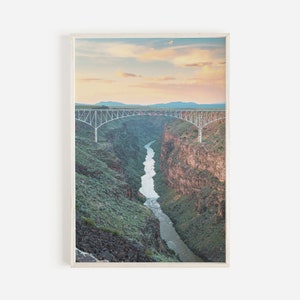 May include: A white bridge spans a deep canyon with a river winding through the bottom. The canyon walls are covered in green vegetation and the sky is a soft pink and orange.
