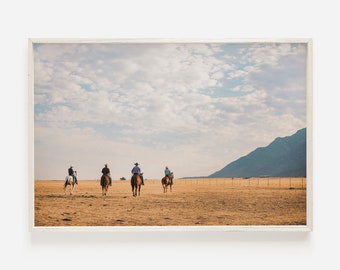 Op de afbeelding: Vijf mensen te paard rijden over een uitgestrekt, droog, bruin veld onder een bewolkte hemel. De ruiters dragen cowboyhoeden en kijken weg van de kijker. In de verte is een bergketen te zien.