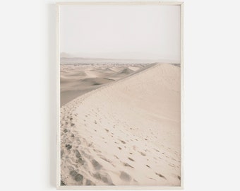 May include: A photograph of a desert landscape with a large sand dune in the foreground. The sand is a light beige color and there are footprints in the sand. The sky is a light blue color and there are mountains in the distance.