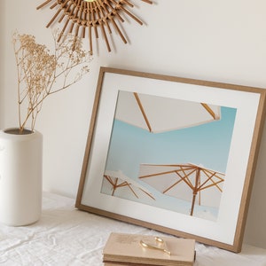 May include: A framed print of a beach scene with three white umbrellas against a blue sky. The frame is made of wood and has a light brown finish.