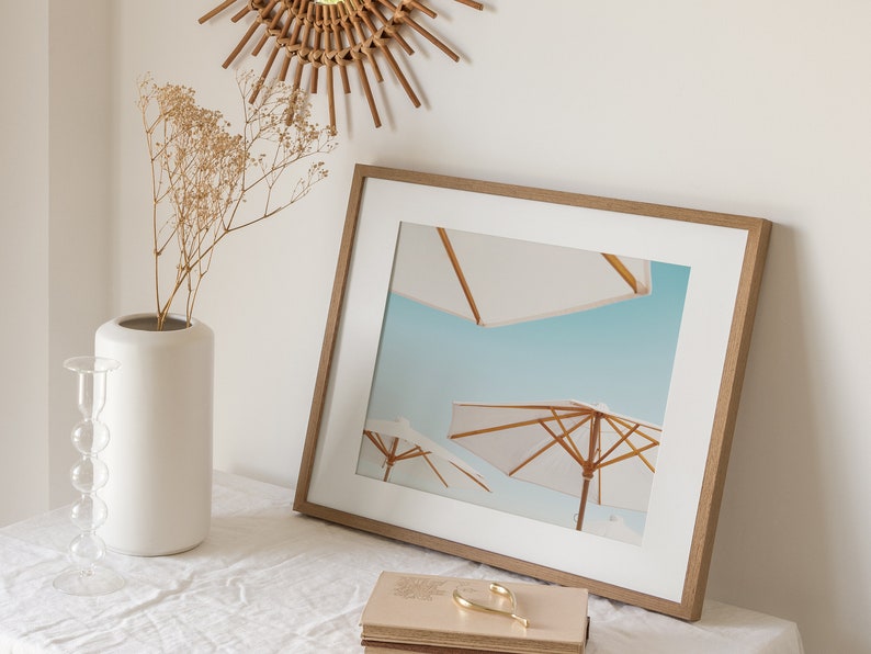 May include: A framed print of a beach scene with three white umbrellas against a blue sky. The frame is made of wood and has a light brown finish.