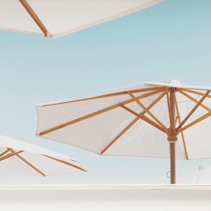 May include: Three white beach umbrellas with wooden frames against a blue sky. The umbrellas are open and the wooden poles are visible.