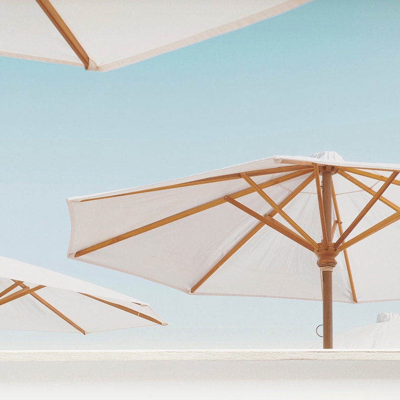 May include: Three white beach umbrellas with wooden frames against a blue sky. The umbrellas are open and the wooden poles are visible.
