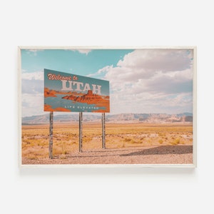 Pode incluir: Um sinal de boas-vindas vintage para Utah, com fundo azul e texto vermelho e branco. O sinal diz "Bem-vindo ao Utah, Life Elevated" e mostra uma imagem cênica de um cânion de rocha vermelha. O sinal está montado em um poste de metal e fica em uma paisagem desértica.