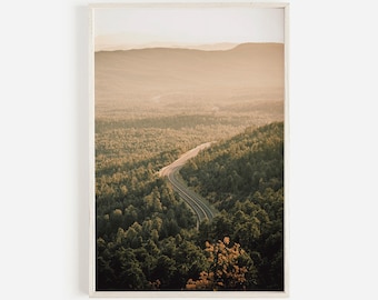 Może przedstawiać: Winding road przez las z pasmem górskim w tle. Droga jest utwardzona, a las gęsty z zielonymi drzewami. Niebo ma delikatny pomarańczowy kolor, co sugeruje zachód lub wschód słońca.