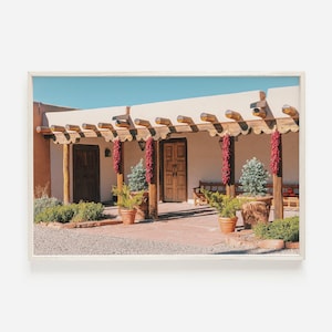 May include: A white adobe building with a terracotta tile roof and a wooden porch. The porch is decorated with red chili peppers hanging from the beams. There are potted plants and a wooden bench on the porch.