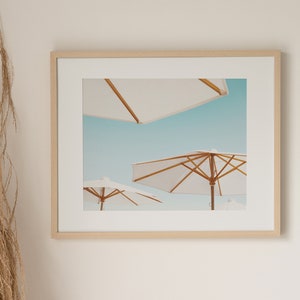 May include: A framed photograph of three white beach umbrellas against a blue sky. The umbrellas are made of white fabric and have wooden frames.