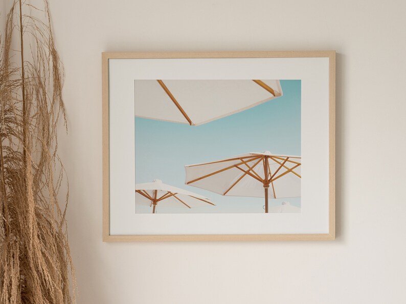 May include: A framed photograph of three white beach umbrellas against a blue sky. The umbrellas are made of white fabric and have wooden frames.