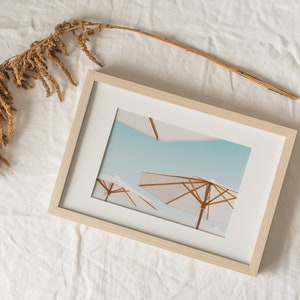 May include: A framed print of a beach scene with two white umbrellas against a blue sky.