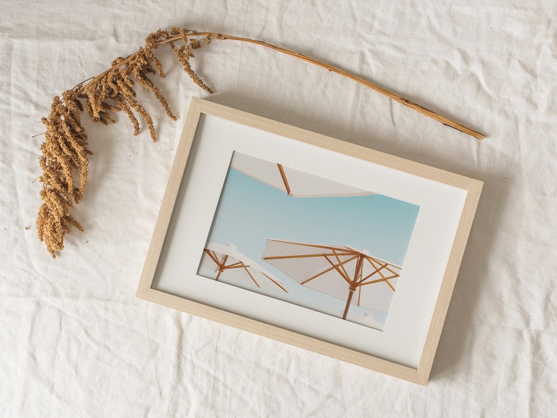 May include: A framed print of a beach scene with two white umbrellas against a blue sky.