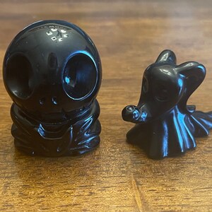 May include: Two black resin figurines, one of a skeleton with a skull head and the other of a ghost with a long flowing robe.