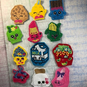 Op de afbeelding: Een set van 12 vingerpoppen van vilt met verschillende voedsel- en drankpersonages. De personages zijn kleurrijk en hebben geborduurde details. De personages omvatten een koekje, een ijslolly, een chocolade reep, een lippenstift, een kolf maïs, een melkpak, een aardbei, een paar schoenen, een broodrooster en een hoge hak.