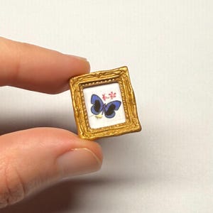 May include: A miniature rectangular picture frame with a metallic gold finish and ornate detailing. The frame contains a small white print featuring a blue butterfly with black markings and a small pink floral accent in the upper corner.