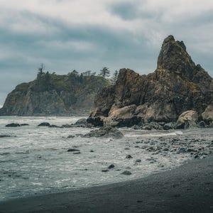 May include: A scenic coastal landscape featuring a dark, rocky beach and turbulent ocean under a cloudy sky. Large, rugged rock formations rise from the shore, with some vegetation. The overall tone is moody and natural.