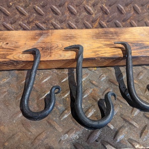 May include: Three black metal hooks with a decorative swirl at the end of each hook. The hooks are mounted on a wooden board.