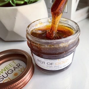 May include: A glass jar filled with a thick, golden brown sugar paste. The jar has a white label with the text "Sweet Sugar Glow" and "Pot of Gold Sugar Paste for all skin types" printed on it. The jar is 4 oz (113.4 g).