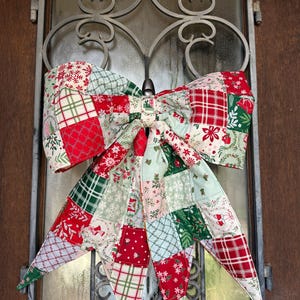 May include: A large decorative bow made of fabric featuring a patchwork design with various holiday-themed patterns in red, green, white, and light green. The fabric squares display motifs including snowflakes, plaid, holly leaves, and small bells.