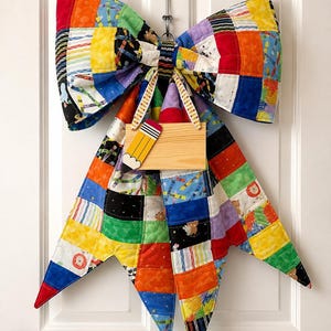 May include: A large, quilted fabric bow featuring a patchwork design of colorful squares with patterns of crayons, bears, and geometric shapes. A small, rectangular wooden sign with a painted pencil graphic hangs from the center of the bow.