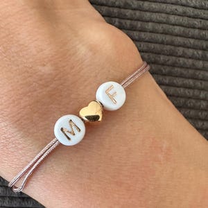 Könnte beinhalten: Ein zartes Armband mit weißen Perlen mit den Buchstaben "M" und "F" in Gold, getrennt durch einen kleinen goldenen Herzanhänger. Das Armband ist an einem dünnen, hellrosa Band befestigt.