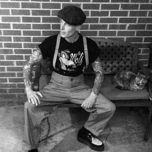 May include: A man wearing a black t-shirt with a cartoon wolf graphic and braces, a black and white newsboy cap, and black and white shoes. He is sitting on a bench with a cat sitting on the bench behind him.