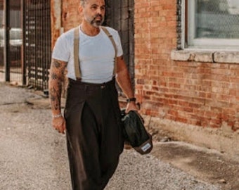 May include: A man wearing a white t-shirt with suspenders and brown wide-leg pants. He is carrying a dark green bag over his shoulder.