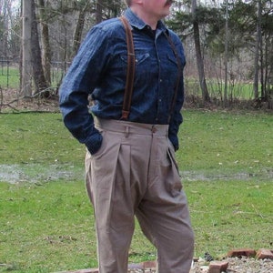 May include: A person wearing a denim shirt with brown suspenders and khaki pleated pants. The person is also wearing brown leather shoes. The outfit is a vintage style.