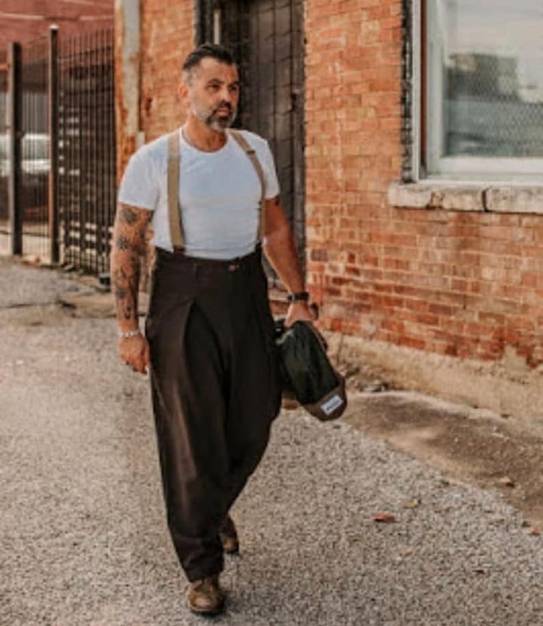 May include: A man wearing a white t-shirt with braces and brown wide-leg trousers. He is carrying a dark green bag over his shoulder.