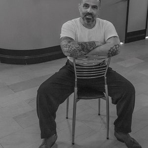 May include: A man with tattoos sits on a metal chair with his arms crossed. He is wearing a white t-shirt, black trousers, and black shoes.