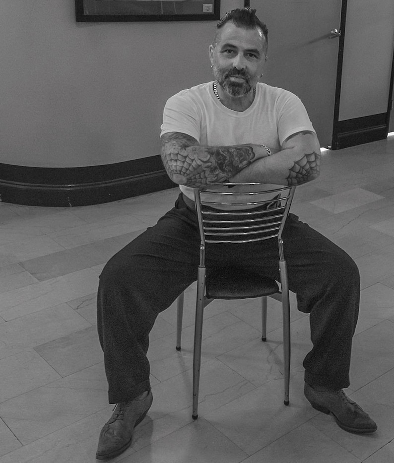 May include: A man with tattoos sits on a metal chair with his arms crossed. He is wearing a white t-shirt, black trousers, and black shoes.