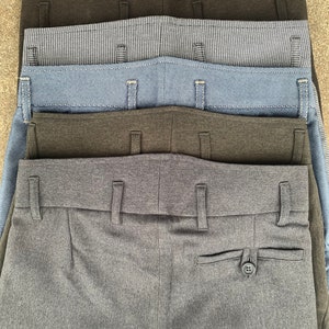 May include: A stack of five pairs of grey, dark grey, blue and black trousers. The trousers are folded and stacked on top of each other. The trousers have a button and a pocket on the back.