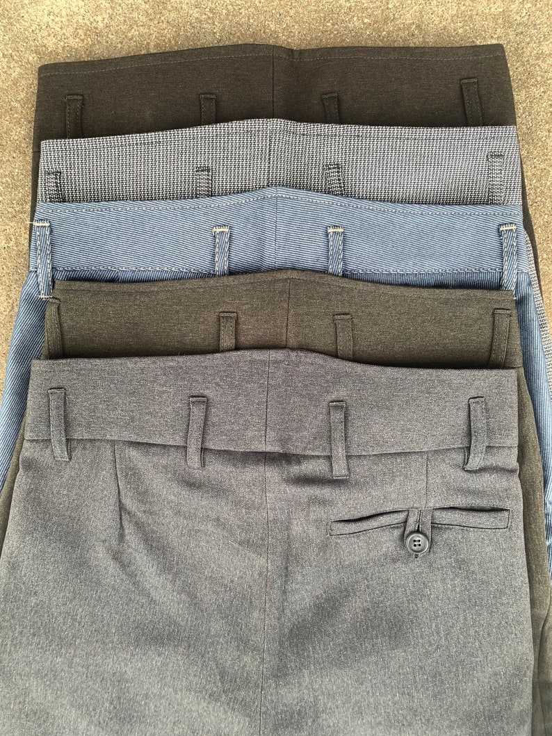May include: A stack of five pairs of grey, dark grey, blue and black trousers. The trousers are folded and stacked on top of each other. The trousers have a button and a pocket on the back.