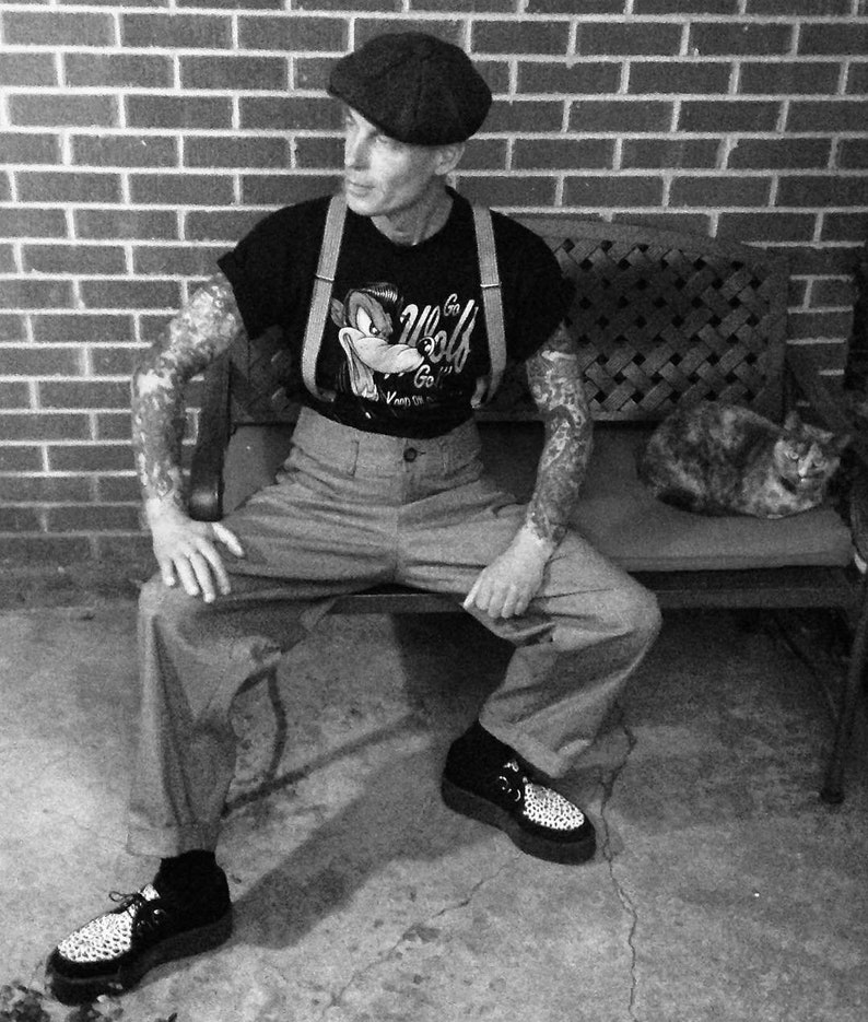 May include: A man wearing a black t-shirt with a cartoon wolf graphic and braces, a black and white newsboy cap, and black and white shoes. He is sitting on a bench with a cat sitting on the bench behind him.