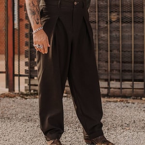 May include: A pair of brown pleated trousers with a button fly and cuffed hems. The trousers are worn with a white shirt and braces.