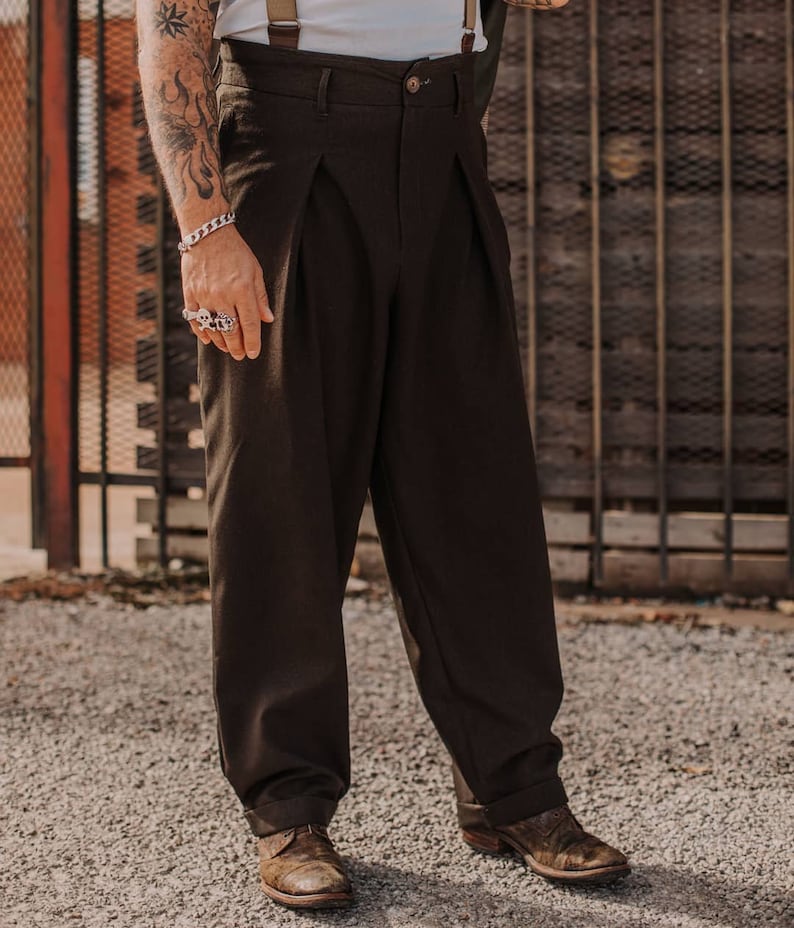 May include: A pair of brown pleated trousers with a button fly and cuffed hems. The trousers are worn with a white shirt and braces.