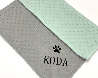 Op de afbeelding: Een grijs en lichtgroen minky-stippen deken met een zwarte pootprint en de naam "KODA" geborduurd erop.