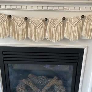 May include: A white macrame garland with wooden beads hangs above a black fireplace. The garland is made of knotted string and has a fringe on the bottom.
