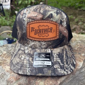 May include: Camouflage trucker hat with a brown leather patch that reads "TC Leather Works." The hat has a black mesh back and a curved brim. A tag on the brim reads "RICHARDSON 112 TRUCKER ADJUSTABLE."