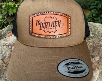 May include: A tan baseball cap with black mesh back panels and a brown leather patch on the front. The patch features the text TC LEATHER WORKS. A circular sticker on the brim reads THE AUTHENTIC YUPOONG SNAPBACK YUPOONG SINCE 1974.