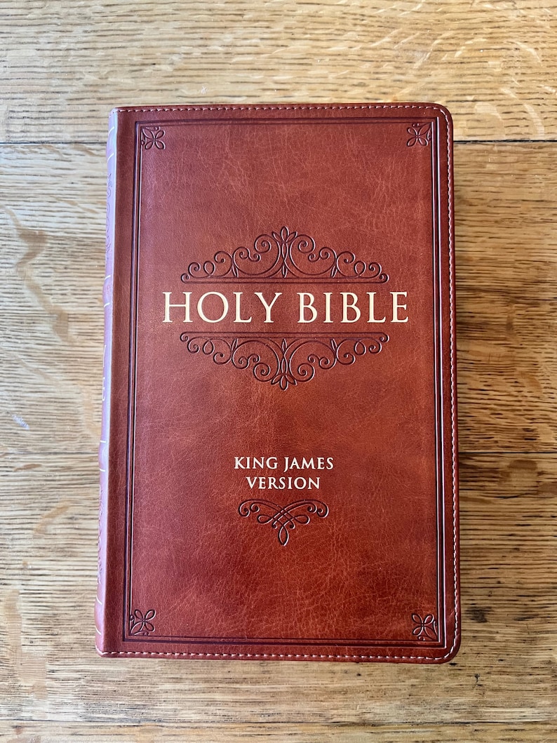 May include: A brown leather-bound Holy Bible, King James Version, with decorative embossed scrollwork and the title in gold lettering. The book is closed and lies on a wooden surface.