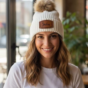 May include: A white knit beanie with a brown faux fur pom-pom on top. The beanie features a brown leather patch with the name "Jordan" stitched on it. The person is wearing a white t-shirt.