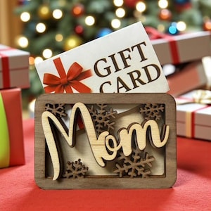 May include: A wooden gift for Mom, featuring the word "Mom" in a cursive font, surrounded by snowflake cutouts. A gift card with a red bow is placed in the wooden frame. The background includes Christmas gifts and a decorated tree.
