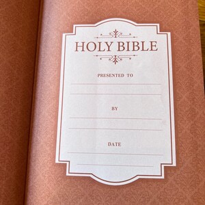 May include: Open Bible with a decorative, patterned cover in shades of peach and burgundy. The inside cover features a white label with the words "HOLY BIBLE" and spaces to write "PRESENTED TO", "BY", and "DATE".