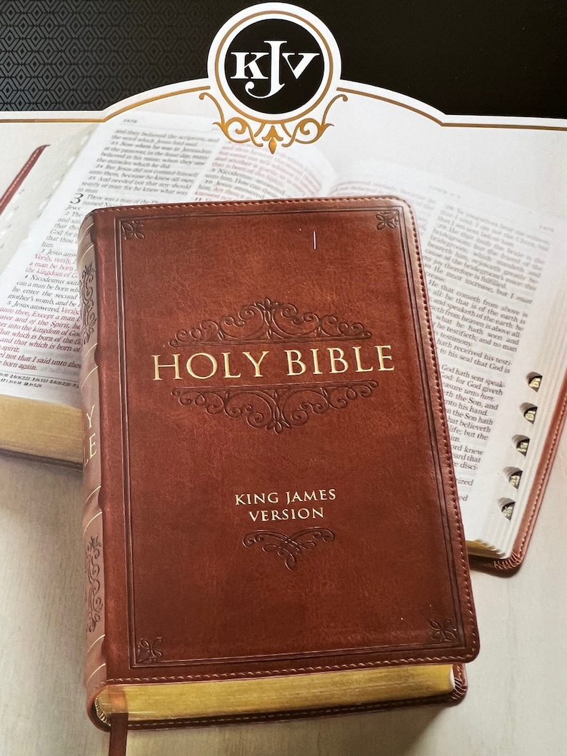 May include: A brown leather-bound Holy Bible, King James Version, with gold-gilded edges. The cover features embossed text and decorative flourishes. The open pages of another Bible are visible in the background.