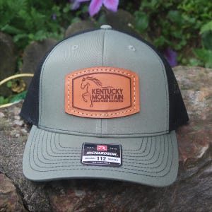 May include: A green and black trucker hat with a leather patch featuring a horse and the words "Kentucky Mountain Saddle Horse Association." The hat has a curved brim and an adjustable strap. The Richardson tag is visible.