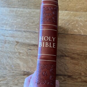 May include: A brown leather-bound Holy Bible with gold lettering. The spine features the text "HOLY BIBLE" and decorative embossed details. The letters "KJV" and "CA" are also visible. The book is held against a wooden surface.