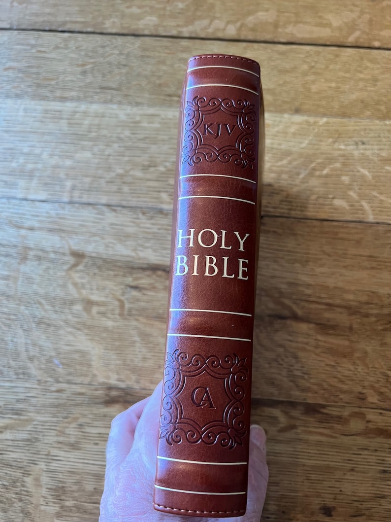 May include: A brown leather-bound Holy Bible with gold lettering. The spine features the text "HOLY BIBLE" and decorative embossed details. The letters "KJV" and "CA" are also visible. The book is held against a wooden surface.