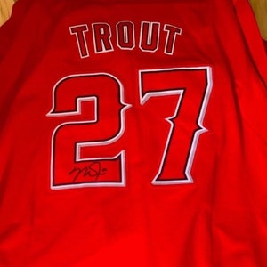 May include: Red baseball jersey with the name "Trout" and the number "27" in white with a black outline. The jersey is signed in black ink.